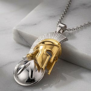Op de afbeelding: Een tweekleurige metalen hanger aan een ketting met een goudkleurige Spartaanse helm, een zilverkleurige kam en een ronde zilverkleurige basis. De hanger hangt aan een fijne zilverkleurige ketting en toont een gedetailleerd ontwerp met gepolijste oppervlakken.