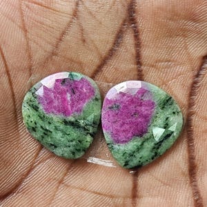 May include: Two faceted, pear-shaped ruby zoisite gemstones featuring a vibrant magenta-pink center surrounded by a mottled, moss-green matrix with dark black inclusions. The stones have a polished, reflective surface with distinct geometric facets.