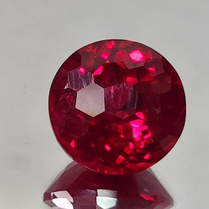 Może przedstawiać: Fasetowany, okrągły, głęboko czerwony kamień szlachetny o błyszczącej, odblaskowej powierzchni. Kamień jest oszlifowany wieloma fasetami, tworząc grę światła i cienia. Kamień jest prawdopodobnie rubinem.