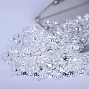Pode incluir: Uma coleção de pequenas pedras preciosas claras e facetadas derramando-se de uma concha prateada. As pedras estão espalhadas sobre uma superfície branca, refletindo a luz e criando um efeito brilhante. A imagem é bem iluminada, destacando a clareza e o brilho das pedras.