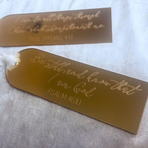 May include: Two gold-colored rectangular tags with rounded tops. The top tag has the text "I can do all things through Christ who strengthens me. PHILIPPIANS 4:13". The bottom tag reads "Be still and know that I am God. PSALM 46:10".