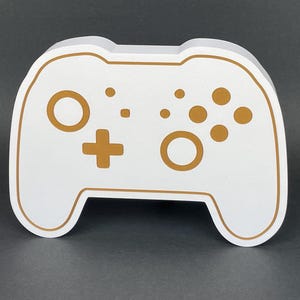 May include: A white foam cutout of a video game controller with gold-colored buttons and a plus sign. The controller is a popular design for gaming consoles.