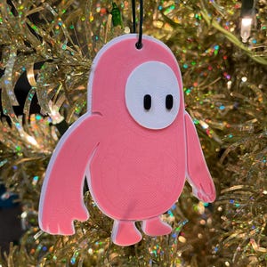 Peut inclure: Une décoration de Noël rose et blanche en forme de personnage du jeu vidéo Fall Guys. La décoration a un visage blanc avec deux yeux noirs.