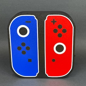 May include: A black, red, and blue Nintendo Switch controller design. The design is made of foam and is a 3D representation of the controller.