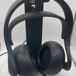 May include: Black over-ear headphones and a matching black stand. The headphones have large, rounded earcups and a curved headband. The stand features a PlayStation logo. The headphones are designed for gaming.