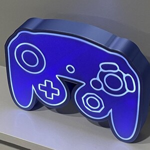 May include: A blue and white GameCube controller shaped night light. The light is on and illuminates the controller in a bright blue colour.