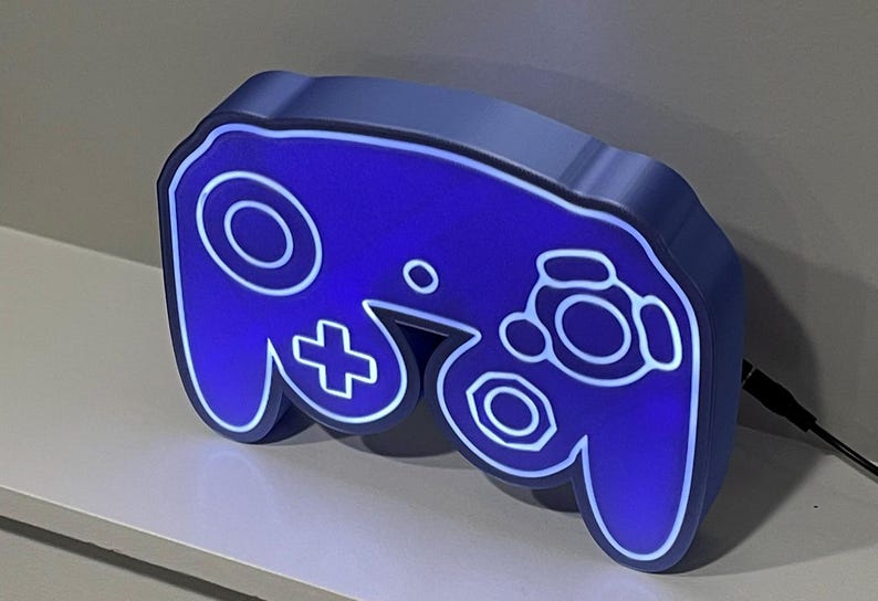 May include: A blue and white GameCube controller shaped night light. The light is on and illuminates the controller in a bright blue colour.