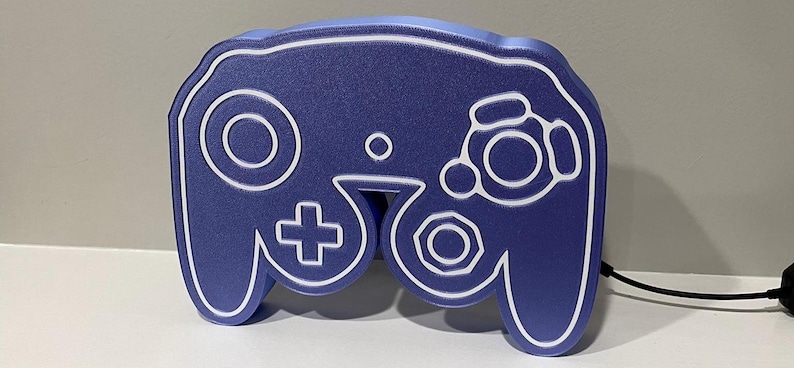 May include: A blue GameCube controller shaped night light with white outlines. The light is plugged in with a black cord.