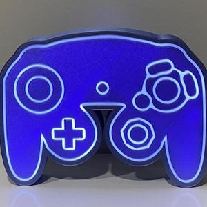 May include: A blue and white GameCube controller shaped night light. The controller is lit up with a white glow.