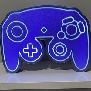 May include: A blue GameCube controller shaped night light with a white outline. The light is on and illuminating the controller.