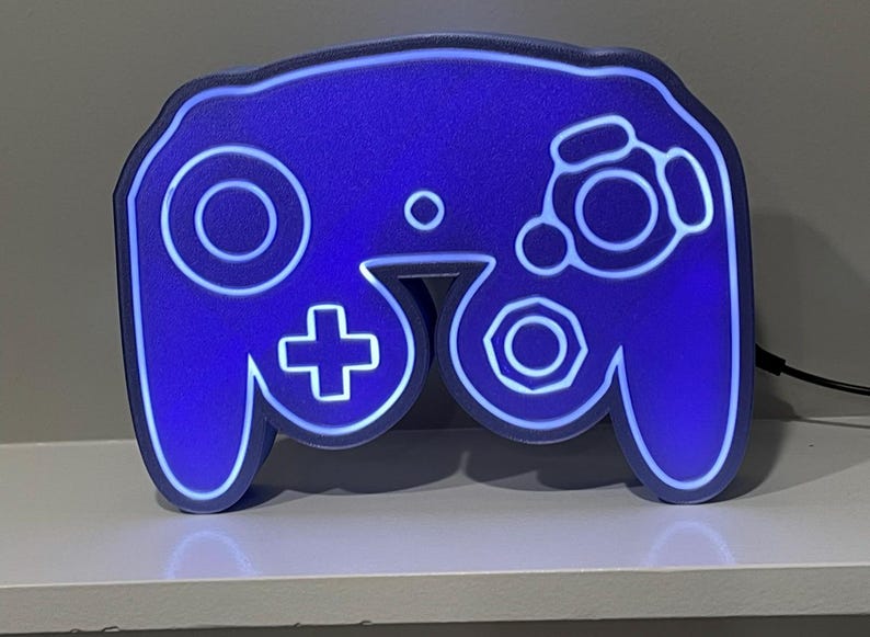 May include: A blue GameCube controller shaped night light with a white outline. The light is on and illuminating the controller.