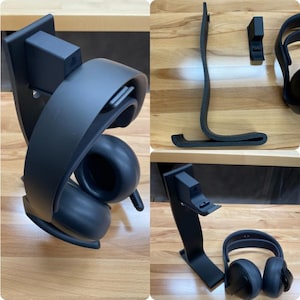 May include: Black headset stand with a curved design, holding a black headset. The stand has a rectangular base and a small, square attachment. The headset is black with rounded earcups.