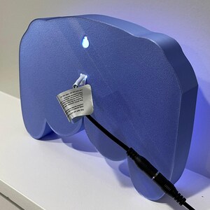 May include: A light blue, 3D printed, controller-shaped night light with a black cord and plug. The light is turned on and emits a blue glow.