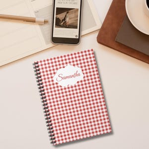 Peut inclure: Un cahier à spirales à carreaux rouges et blancs avec le nom "Samantha" sur une étiquette blanche. Un stylo, un téléphone, un agenda et une tasse de café sont également visibles.