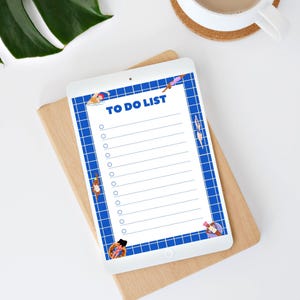 Könnte beinhalten: Ein weißes Tablet mit einer "TO DO LIST" mit blauem Gitterrand und Illustrationen von schwimmenden Personen. Das Tablet liegt auf einer Holzoberfläche, im Hintergrund eine Kaffeetasse und eine Pflanze.