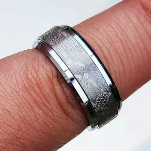 May include: A silver tungsten carbide ring with a bevelled edge and an etched design of sea turtles.