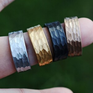 May include: Four metal rings with a hammered texture, arranged in a row. The rings feature distinct finishes including a silver-tone, a bright gold-tone, a matte black, and a warm copper-tone, each showcasing a reflective, uneven surface pattern.