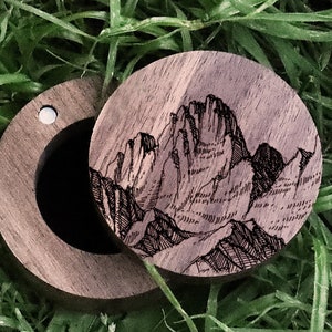 May include: A wooden ring box with a hinged lid. The lid features a detailed engraved design of a mountain range. The box is made of dark wood and has a smooth finish.