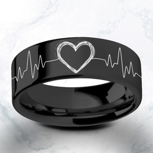 以下が含まれることがあります： 白い心臓と心拍のデザインが施された黒いタングステンカーバイドリング。リングには心拍線と心臓が刻まれています。