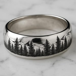 Puede incluir: Un anillo de plata con un diseño grabado de una cordillera y árboles. El anillo es una banda ancha con un borde redondeado.