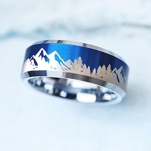 May include: A silver ring with a blue band featuring a mountain and forest design. The ring has a polished finish and a detailed landscape engraving. The ring's design is a nature-inspired accessory.