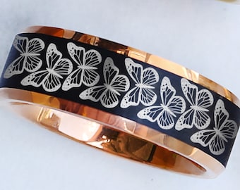 May include: A ring featuring a black band with a repeating laser-engraved butterfly pattern and a polished rose gold interior. The design showcases multiple butterflies arranged in a continuous row around the circumference of the metal band.