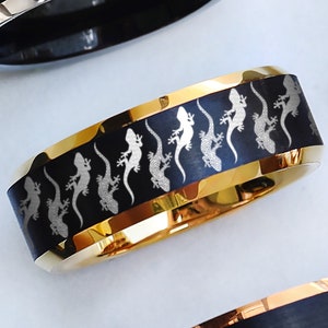 May include: A black and gold tungsten carbide ring with a pattern of white gecko silhouettes. The ring has a brushed finish and a polished edge.