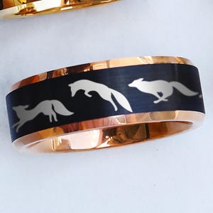 May include: A black and rose gold ring with a design of three white foxes running in a line. The ring is made of tungsten carbide and has a polished finish.