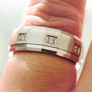 May include: A silver-toned ring with a geometric design, featuring small square diamond accents. The ring is worn on a finger, showcasing its polished and textured surfaces. A modern piece of jewelry.