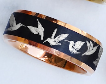 May include: Rose gold ring with a black band featuring engraved white hummingbird designs. The ring has a gold-colored interior and exterior trim. The design is repeated around the band. The ring is shown on a white surface.
