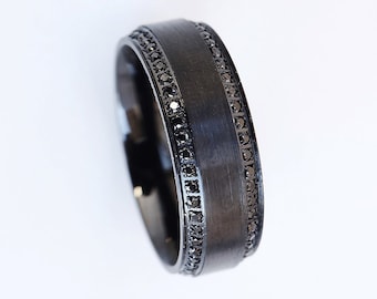May include: A black metal ring featuring a wide, brushed-finish centre band flanked by two narrow channels. Each channel is set with a continuous row of small, dark, faceted stones that provide a subtle sparkle against the dark metal surface.