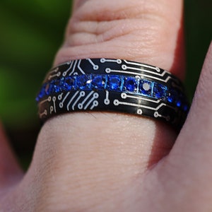 May include: A black ring with a circuit board design and a row of blue gemstones.