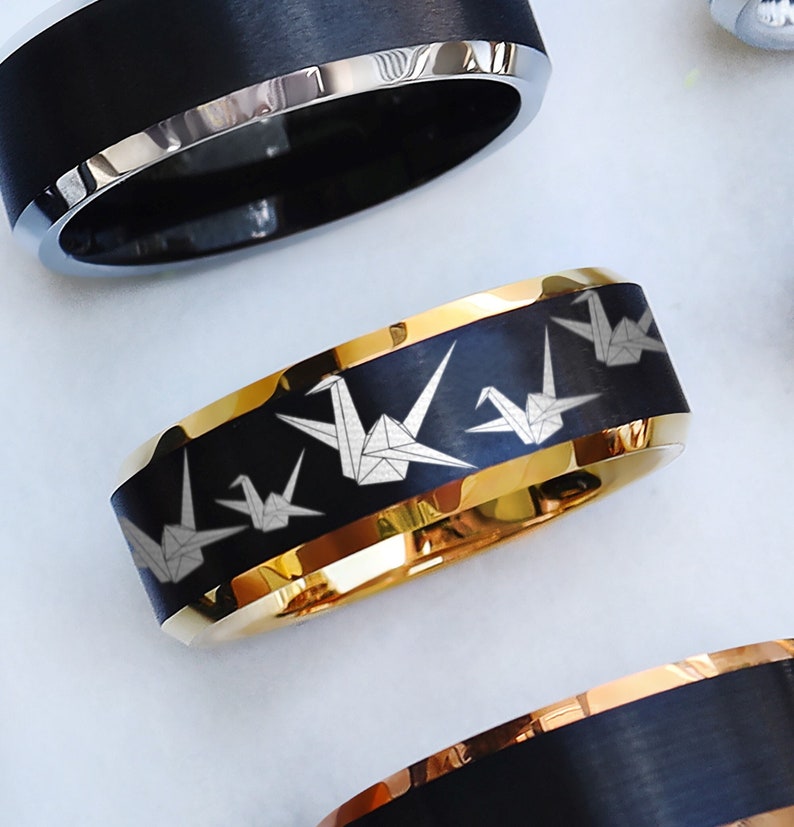 May include: Close-up of several rings. One ring features a black band with a gold interior and trim, and a pattern of white origami cranes. Other rings have black bands with silver or gold trim.