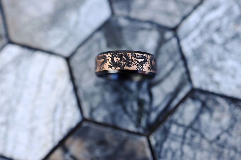 K&ouml;nnte beinhalten: Ein schwarzer und ros&eacute;goldener Ring mit einem Drachenmotiv. Der Ring liegt auf einer grauen und wei&szlig;en gekachelten Oberfl&auml;che.