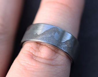 May include: A silver-colored ring with a brushed finish. The ring features an engraved design of a wave. The ring is shown on a finger.