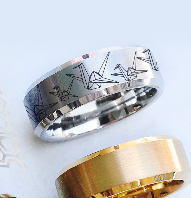 May include: A silver-colored ring with a brushed finish and a band featuring a repeating pattern of origami cranes. The ring has a polished interior and a beveled edge. A gold-colored ring with a brushed finish is partially visible below.