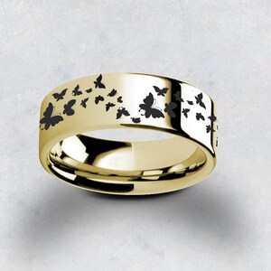Pu&ograve; includere: Un anello a fascia d'oro con un design di farfalle nere. Le farfalle sono disposte in un motivo attorno alla fascia.