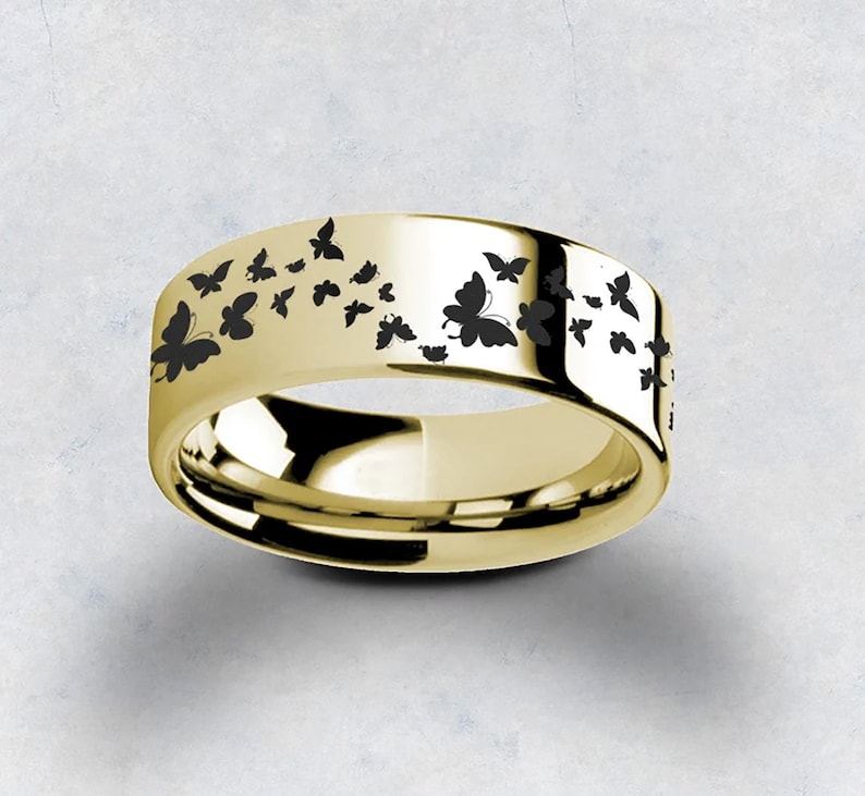 Pu&ograve; includere: Un anello a fascia d'oro con un design di farfalle nere. Le farfalle sono disposte in un motivo attorno alla fascia.