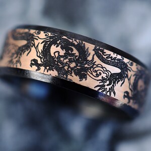 K&ouml;nnte beinhalten: Ein schwarzer und ros&eacute;goldener Ring mit einem gravierten Drachenmuster auf der Band.