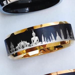 May include: A gold-toned band ring featuring a black center inlay with a laser-engraved white graphic depicting a seated Buddha figure and various architectural structures, including temple spires and buildings, wrapping around the entire circumference.