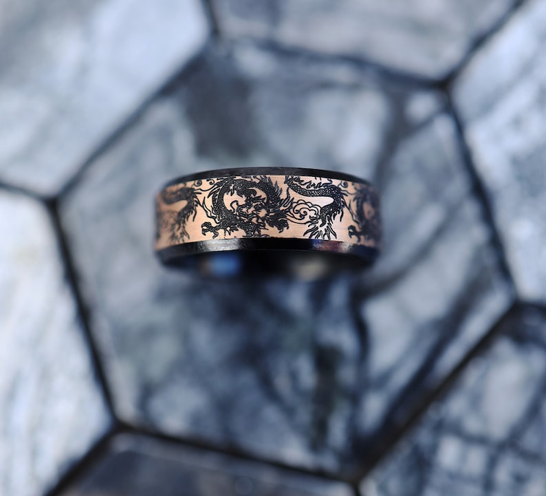 K&ouml;nnte beinhalten: Ein schwarzer Ring mit einer ros&eacute;goldenen Banderole, die mit einem Drachenmotiv verziert ist.