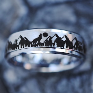 May include: A silver ring featuring a black silhouette of a wolf howling at the moon in front of a mountain range. The ring is engraved with a detailed design.