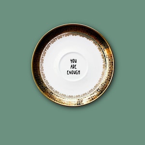 Könnte beinhalten: Eine weiße Untertasse aus Keramik mit einem breiten, dunkelgoldenen Rand und einem komplexen Wirbelmuster. In der Mitte der Untertasse befindet sich der schwarze Schriftzug "YOU ARE ENOUGH" in einer einfachen Schriftart, zentriert in der Vertiefung.