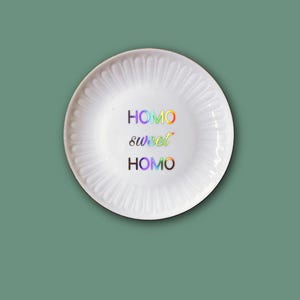 Könnte beinhalten: Ein weißer Keramikt weißer Keramikt weißer Keramikteller mit gewelltem Rand und glänzender Oberfläche. In der Mitte des Tellers befindet sich der Schriftzug "HOMO sweet HOMO", der in einem lebendigen, mehrfarbigen holografischen Verlauf gedruckt ist.