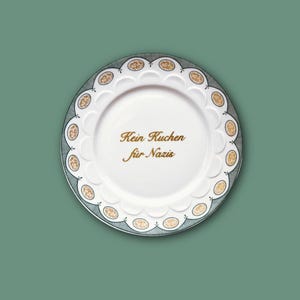Könnte beinhalten: Ein weißer Keramikteller mit gewelltem Rand, verziert mit einer Bordüre aus kleinen goldfarbenen Ovalen und feinen schwarzen Linien. In der Mitte des Tellers befindet sich der Schriftzug "Kein Kuchen für Nazis" in goldfarbener Schreibschrift.