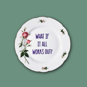 Könnte beinhalten: Ein weißer Keramikteller mit gewelltem Rand, verziert mit einem Blumenmuster aus rosa Rosen und grünen Blättern auf der linken Seite sowie kleinen Rosenknospen am Rand. In der Mitte steht der violette Text, WHAT IF IT ALL WORKS OUT?