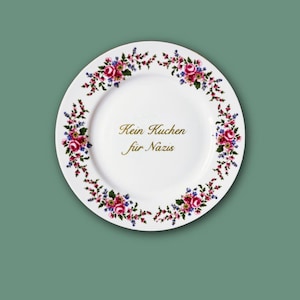 Könnte beinhalten: Ein weißer Keramikteller mit einem floralen Rand aus rosa Rosen und kleinen blauen Blumen. In der Mitte des Tellers befindet sich der goldfarbene Schriftzug "Kein Kuchen für Nazis" in kursiver Schrift, der in zwei Zeilen auf der Fläche steht.
