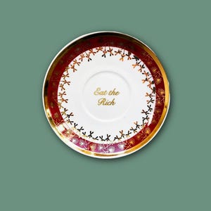 Könnte beinhalten: Eine runde Untertasse aus weißer Keramik mit einem dunkelroten Rand und einem goldenen Pusteblumenmuster. In der Mitte der Untertasse steht der Schriftzug Eat the Rich in goldener Farbe. Der Rand ist mit einem dünnen Goldstreifen verziert.