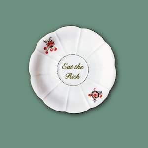 Könnte beinhalten: Eine weiße, gewellte Untertasse aus Keramik mit dem Schriftzug Eat the Rich in goldener Schrift in der Mitte. Der Teller ist mit zwei kleinen, symmetrischen Blumenmotiven in Rot, Schwarz und Weiß an gegenüberliegenden Seiten des Randes verziert.