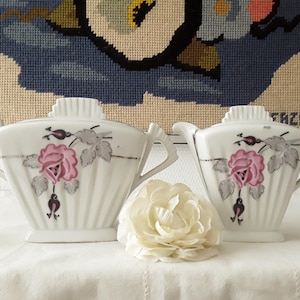 Peut inclure: Service à thé en porcelaine blanche avec un motif de rose rose et de feuilles grises. L'ensemble comprend une théière et un sucrier. Une fleur blanche est devant le service à thé. Le fond présente une tapisserie.